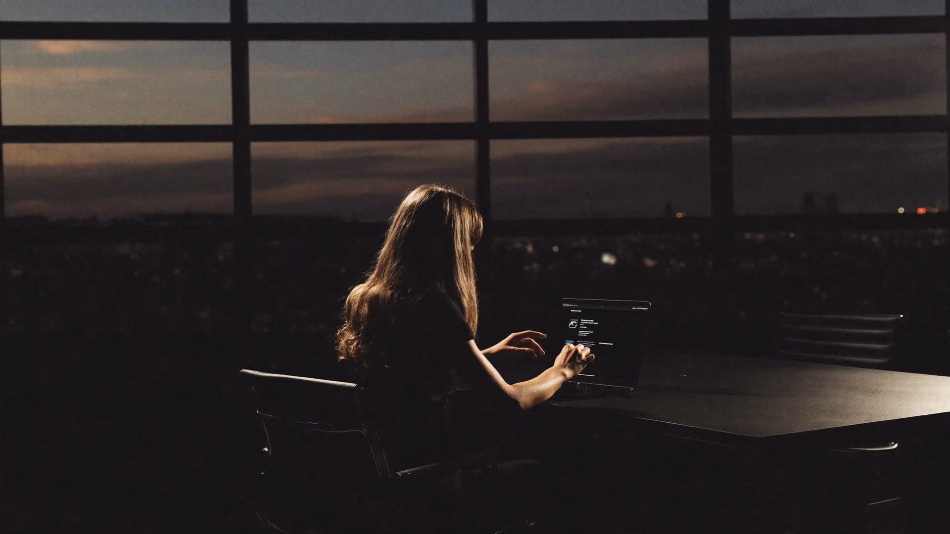 A dimly lit office at dusk, one person still working by the glow of a laptop
